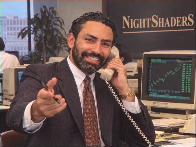 An image from the film NightShaders, showing a smiling bearded man in a pinstripe suit talking on a telephone with stock charts on a computer monitor behind him.