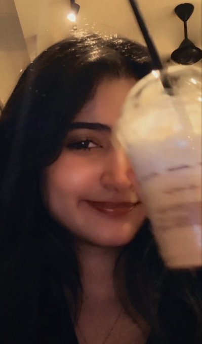 A woman with long dark hair smiles softly while posing next to a glass containing a white frozen drink topped with whipped cream and a black straw.