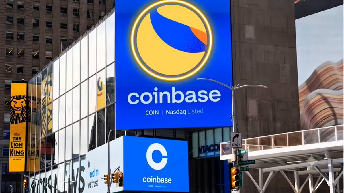 The image shows a city street with a large blue coinbase sign, which is located near a tall building. The sign has an orange border and features a yellow circle with a blue line through it. Below the sign, there are two smaller signs that read "coinbase" and "LUNC". These signs are placed on top of a building, possibly indicating the presence of a cryptocurrency exchange or news outlet related to coinbase. The image also contains text that reads "BREAKING NEWS $LUNC is live in time square New York", which suggests that there might be breaking news or an important event happening at this location.