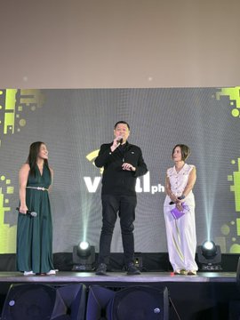 The image features a man standing on stage holding a microphone while two women stand behind him, both wearing dresses. They appear to be part of an event or performance related to the Viral.PH campaign that aims to promote emerging technologies like blockchain. The backdrop is a large screen displaying the Viral.PH logo and text, indicating the theme and purpose of the gathering.