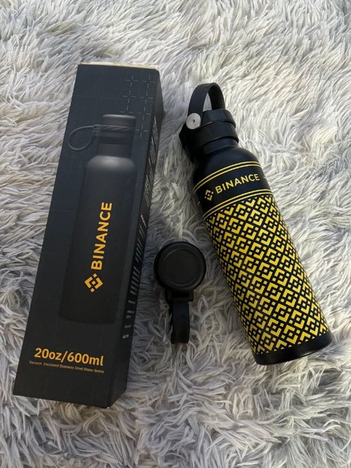 A black Binance-branded insulated stainless steel water bottle (20oz/600ml) with a yellow geometric pattern sits next to its packaging box and a separate lid on a fluffy white rug, showcasing the sleek merchandise item referenced in the caption as a thoughtful community gift from Binance.