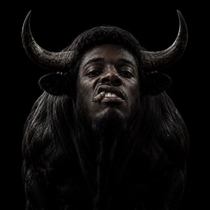 A dark, dramatic portrait of a person with bull horns, a nose ring, and long dark hair, set against a black background.