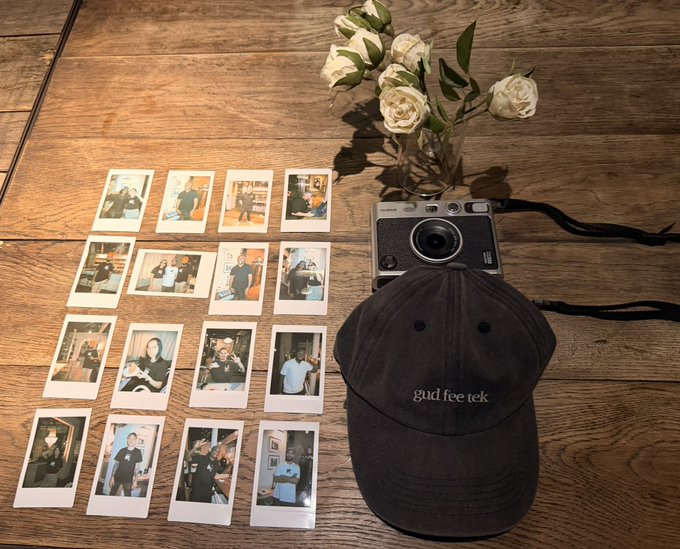 A flat lay on a wooden table featuring a vintage camera, a vase of white roses, a black 'gud feet' cap, and a grid of sixteen polaroid photos.