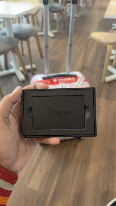 A hand holds up a black 'Tangem' branded card in front of a red suitcase in a room with stools and tables visible in the distance.