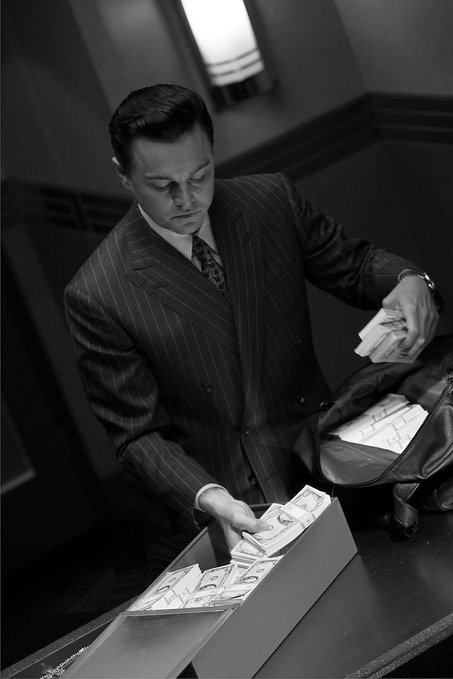 A black-and-white photograph of a man in a pinstripe suit packing stacks of US dollar bills into a bag and a box.