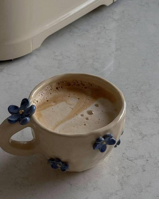A close-up of a ceramic mug with blue flower decorations filled with a frothy coffee drink, sitting on a marble countertop next to a white appliance.