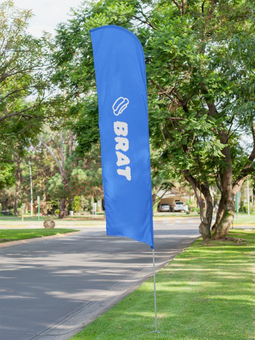 A tall blue advertising flag displaying "BRAT" with a bread icon stands beside a paved path in a tree-lined park area.