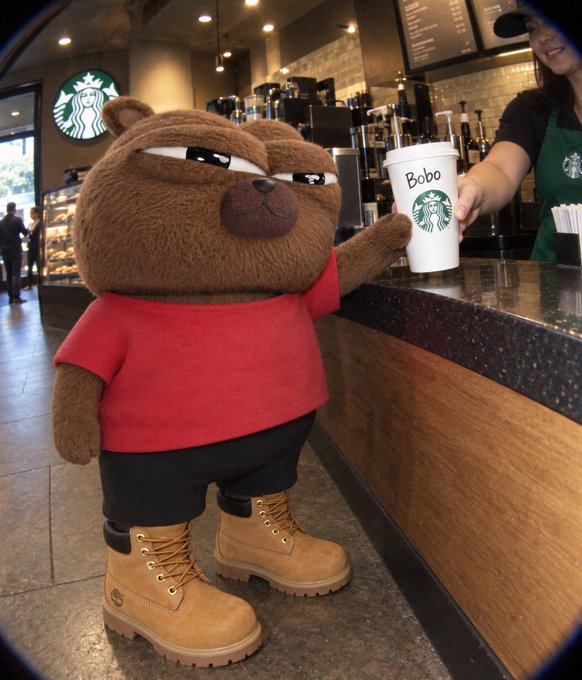 A 3D-rendered brown bear character wearing a red t-shirt, black shorts, and tan Timberland boots stands at a Starbucks counter receiving a white cup labeled 'Bobo' from a barista.