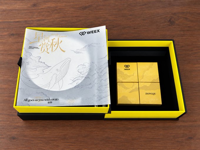 An open yellow WEEX branded gift box featuring a silver booklet with whale designs and the text 'All goes as you wish (如愿)' alongside four smaller yellow boxes.