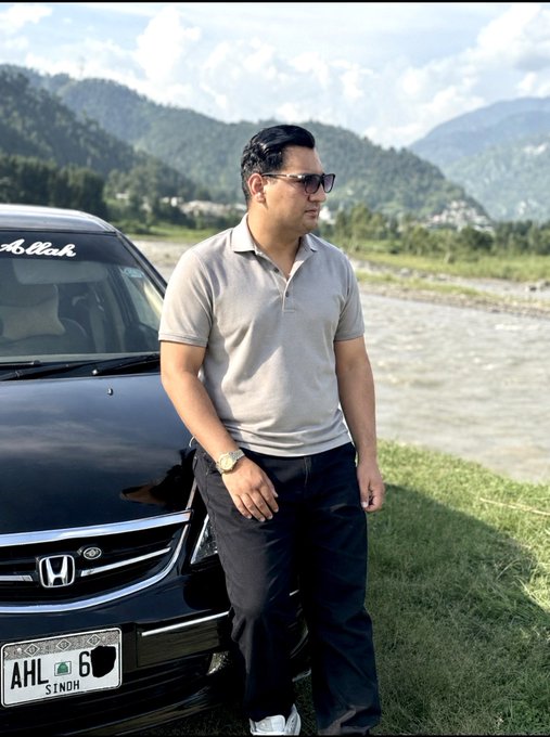 A man in sunglasses and a gray polo shirt leans against his black Honda car, with the windshield decal reading "Ashish" and license plate "AHL 6 SINOH," as he takes a peaceful break by the riverside with mountains in the background. The serene outdoor setting reflects his post about stepping away from risky crypto trades to relax and reset his mindset with his wife.