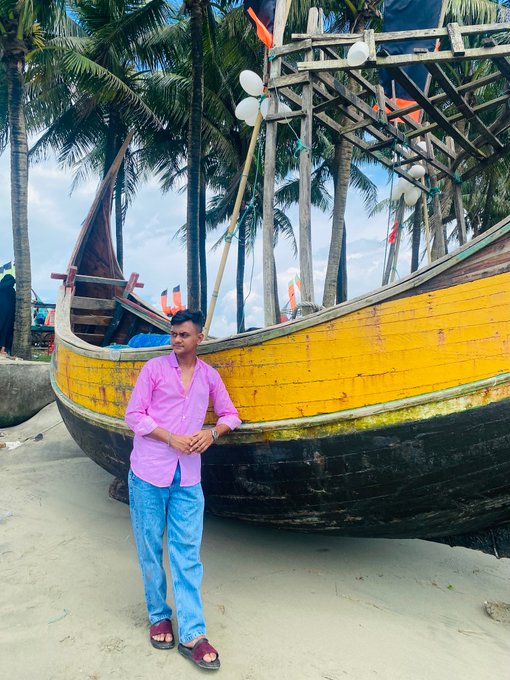 A man wearing a pink button-up shirt, light blue jeans, and dark sandals stands posing on a sandy beach in front of a large traditional wooden fishing boat painted in vibrant yellow and black, surrounded by tall palm trees under a partly cloudy sky.
