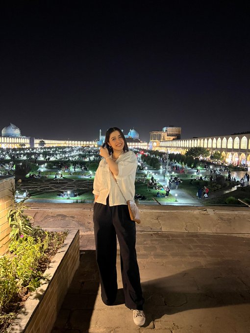 A woman smiles softly at the camera while standing on a rooftop terrace at night, with the illuminated Naqsh-e Jahan Square in Isfahan, Iran beautifully glowing behind her. The serene nighttime backdrop complements the post's hopeful message of manifesting a sell-out for her artwork collection on objkt, capturing a moment of quiet confidence as she shares her creative pieces with the world.