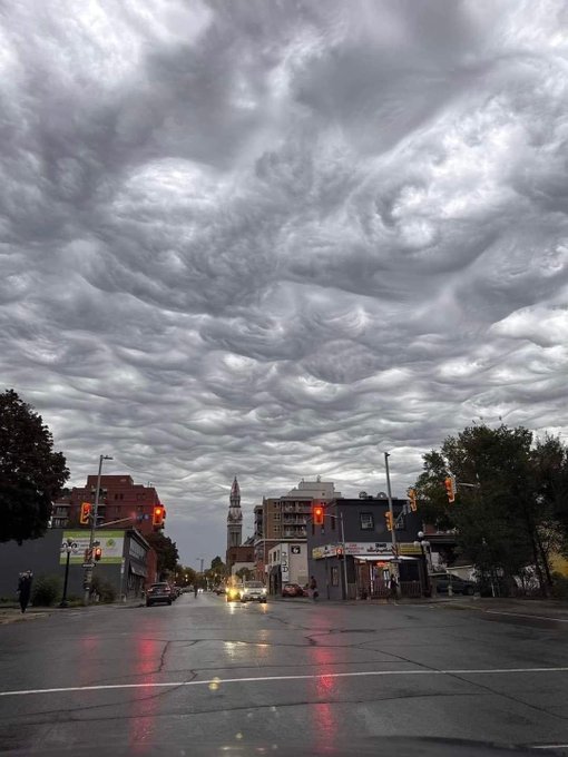 The image shows an urban street scene with buildings lining both sides of the road. The sky above has large clouds, creating a dramatic backdrop for the cityscape below. There are several cars parked along the side of the road, as well as traffic lights visible at various points in the scene. A person can be seen walking on the sidewalk near one of the buildings.