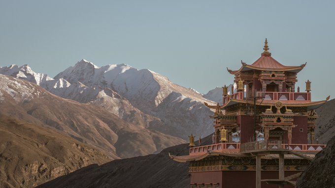 A scenic photograph of an ornate red and gold Tibetan-style pagoda set against a backdrop of snow-capped Himalayan mountains under a clear sky.