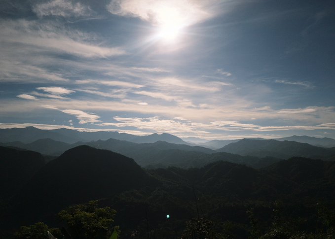 A serene mountain landscape stretches into the distance beneath a bright sun and scattered clouds, evoking a sense of calm and escape. The image captures the speaker's brief break from their usual talking-head videos, with rolling, forested hills and hazy peaks suggesting a moment of "touching grass" before returning to their content.
