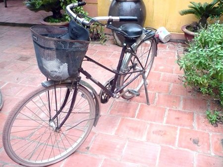 An old black bicycle with a front basket sits on a red-tiled patio near potted plants and a yellow wall.