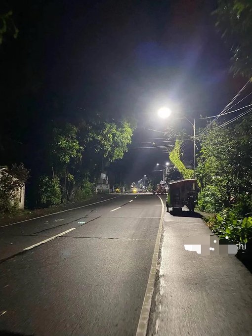 The image shows an empty street at night with a car driving down the road, illuminated by a street light. The scene appears to be captured during the day when the sun sets, creating a dark atmosphere. There are no people or other vehicles visible in the photo. The focus of the image is on the car and its journey along the deserted street under the glow of the street light.