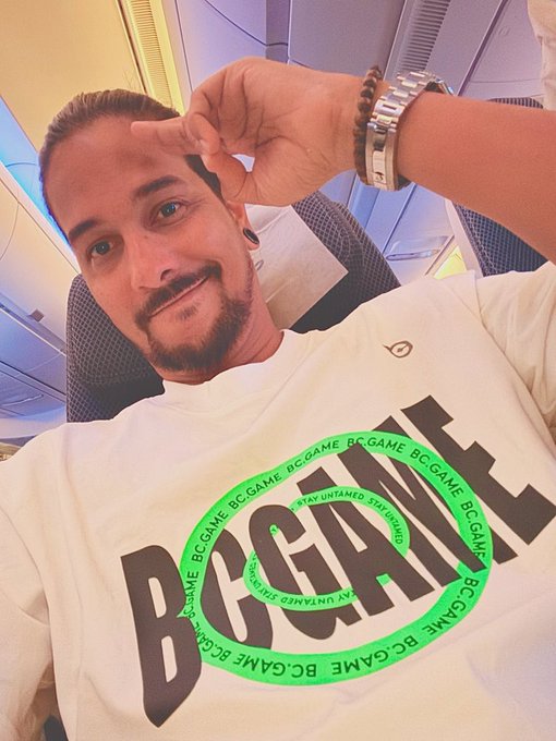 A man on an airplane smiles while resting his hand near his forehead, wearing a white t-shirt featuring the "BC.GAME" logo in bold black letters within a green circular design that reads "STAY UNTAMED." The image complements the "Gm Fam" post by showcasing his casual, branded crypto-casino apparel in a relaxed travel setting.