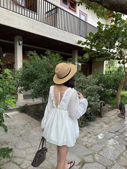 A photo of a woman from behind wearing a straw hat and a white summer dress, walking through a garden courtyard with stone pavement and a two-story building with wooden railings.