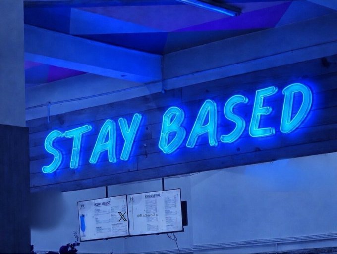 A glowing blue neon "STAY BASED" sign illuminates the wall of what appears to be a venue, with smaller posters below referencing B20 and Base. The image ties to the announcement of B20 launching on the Base ecosystem, with the "STAY BASED" message serving as a fitting rallying cry for the community milestone.