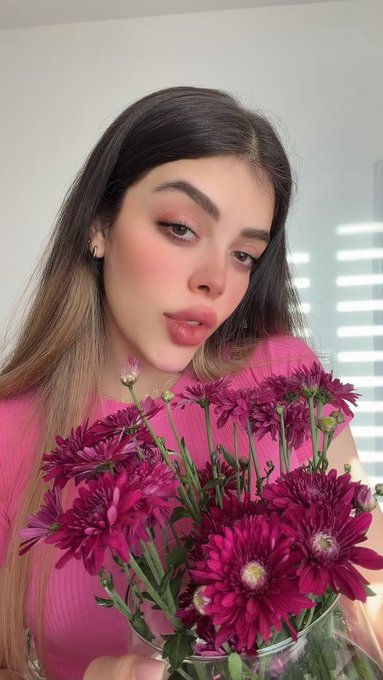 A young woman with long brown hair wearing a pink top poses for a selfie while holding a bouquet of bright pink chrysanthemums.