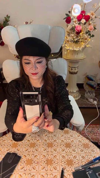 A woman wearing a black beret and a face veil, looking down at a white smartphone she is holding.
