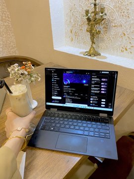 A person wearing gold bracelets holds an iced coffee next to a laptop displaying an audio editing software, set on a wooden table near a decorative candelabra.