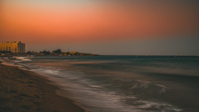 A serene beach scene at sunset with orange sky, waves rolling onto the shore, and buildings visible along the coastline in the distance.