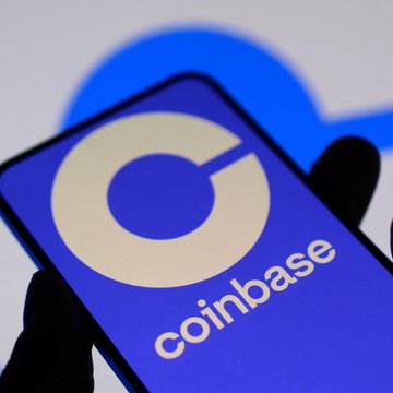 A gloved hand holding a smartphone displaying the Coinbase logo and name in white text on a blue background.