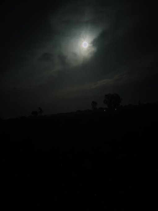 A nighttime photograph of a bright full moon partially obscured by dark clouds, with silhouetted trees visible along the horizon.