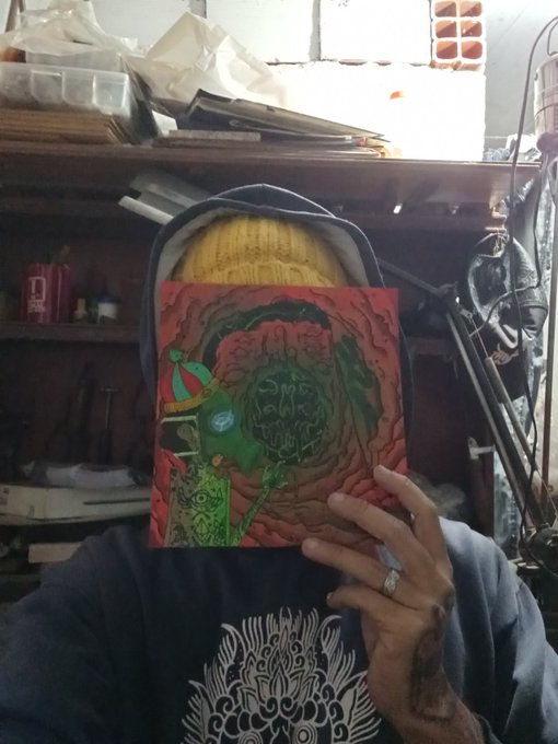 A person wearing a hoodie holds up a vibrant red and green artwork featuring a tentacled creature face, standing in what appears to be a cluttered workshop or studio space.