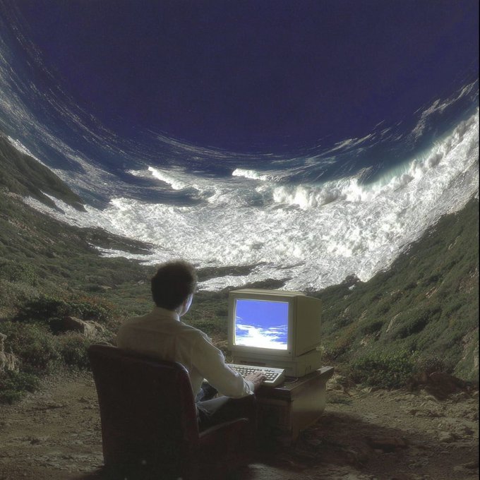 A surreal digital composite shows a person in a white shirt sitting at a vintage CRT computer in a chair outdoors, with massive ocean waves towering above them against a dark blue sky.