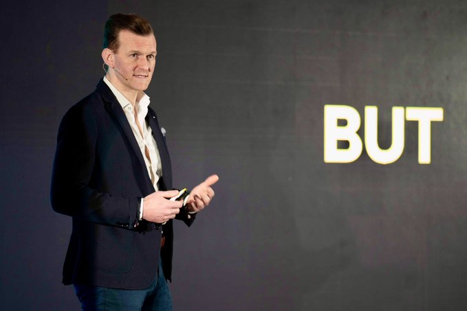 A man in a dark blazer gives a presentation on stage, gesturing toward a large screen displaying the word "BUT" in bold white letters. The image captures a moment from his keynote at the World Economic Forum 2026 Web3 Hub, where the prominent "BUT" text visually emphasizes the counterpoints or challenges he raised about institutional blockchain adoption.
