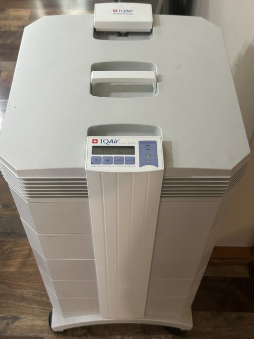 A white IQAir air purifier is pictured on a wooden floor, featuring the brand logo on the top and front panels along with a digital display and the text "Ion Purification + Electrostatic Capture."