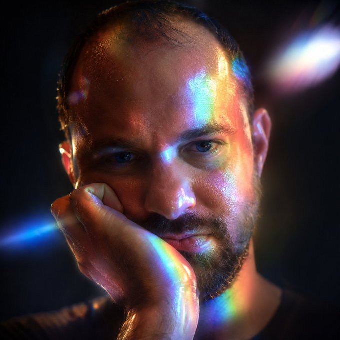 A close-up portrait of a bearded man resting his chin on his hand, illuminated by colorful prismatic lighting against a dark background.