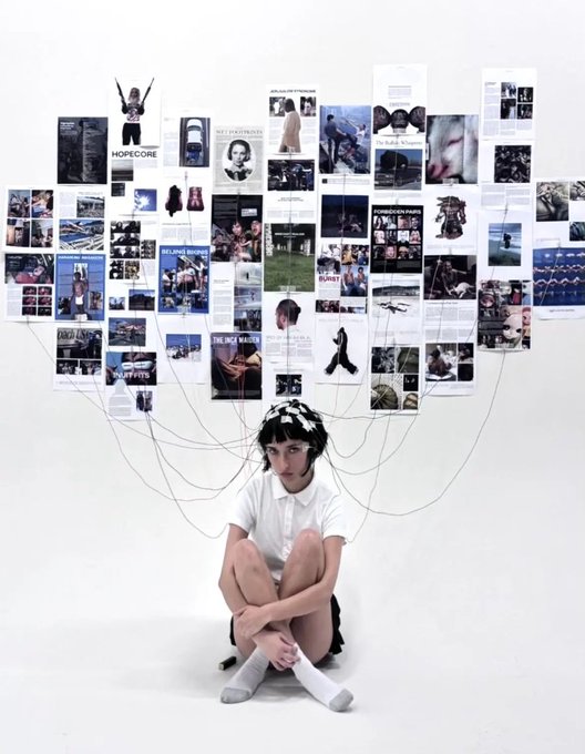 An artistic shot of a girl with a floral crown sitting on the floor surrounded by an extensive wall collage of magazine clippings and photos connected by strings to her head.