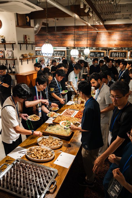 A crowded room at the Injective Ecosystem Meetup in Tokyo shows attendees serving themselves from a long buffet table laden with various dishes, reflecting the post's emphasis on industry professionals gathering to discuss practical blockchain deployment rather than its theoretical value.