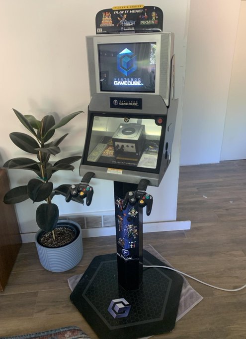 A custom-built Nintendo GameCube arcade kiosk stands in a room, featuring a top-mounted screen displaying the "Nintendo GameCube" logo, a central display case housing an actual GameCube console, and two GameCube controllers mounted on a decorated pedestal next to a potted rubber plant. The header marquee reads "PLAY IT HERE!" and showcases game artwork including Pikmin.