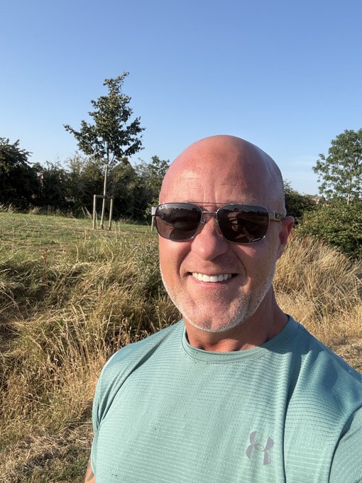 Selfie of a smiling bald man wearing sunglasses and a teal Under Armour shirt standing in a grassy field with trees.