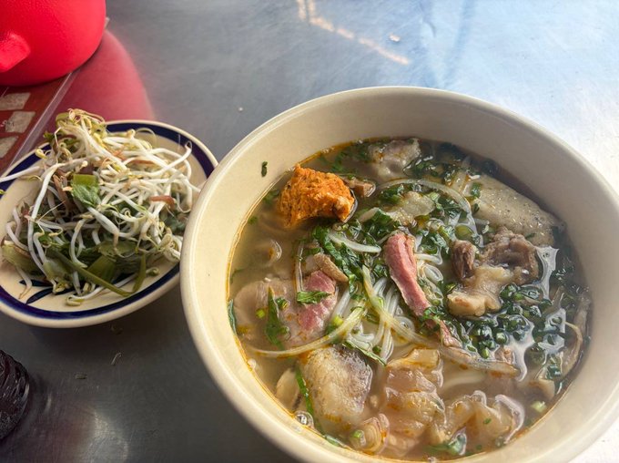 A large bowl of Vietnamese noodle soup with meat and greens in a clear broth, served alongside a smaller plate of fresh bean sprouts and herbs on a metal table.