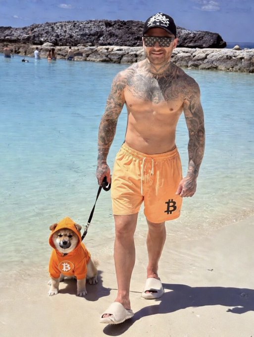 A heavily tattooed shirtless man wearing orange Bitcoin-themed swim trunks, a black Bitcoin cap, sunglasses, and slides, walking his corgi on a leash at a beach; the dog is dressed in a matching orange Bitcoin hoodie.