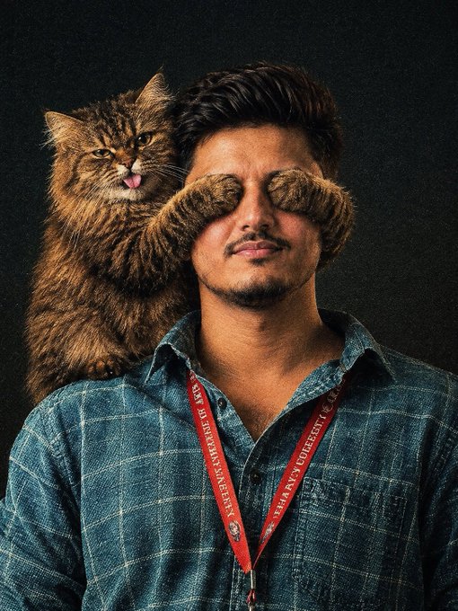 A portrait photo of a man wearing a denim shirt and a red lanyard, with a fluffy brown cat perched on his shoulder appearing to cover his eyes with its paws like a mustache.