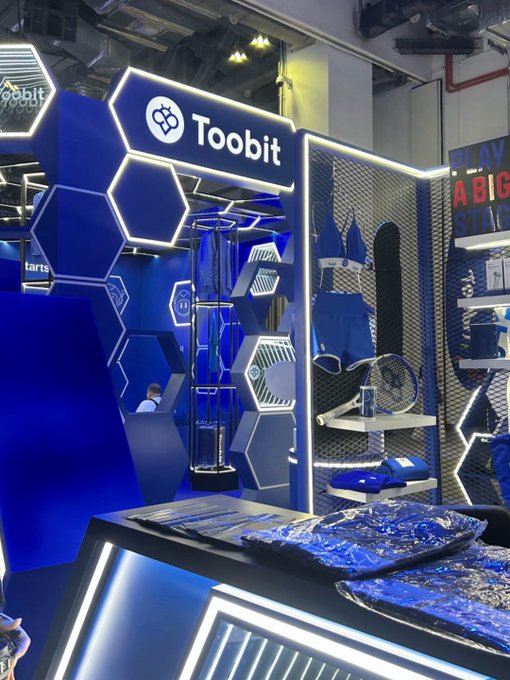 A trade show booth for the cryptocurrency exchange Toobit features a blue and white hexagonal design with merchandise displayed on the right.