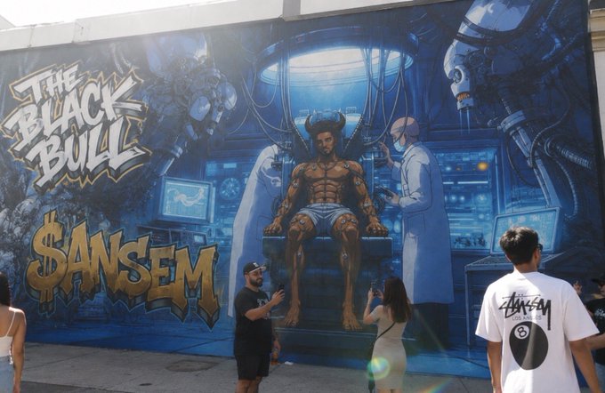 A vibrant blue mural on a Los Angeles wall depicts a horned, tattooed man seated in a sci-fi chair while a scientist observes, labeled "The Black Bull" and "$ANSEM." A few pedestrians, including one wearing a Stüssy shirt, stop to admire and photograph the street art while walking around the city.