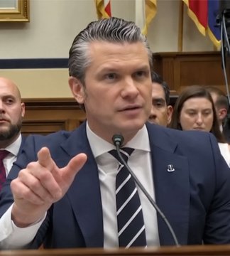 A man in a navy blue suit with a striped tie speaks into a microphone and gestures with his hand during what appears to be a formal hearing.