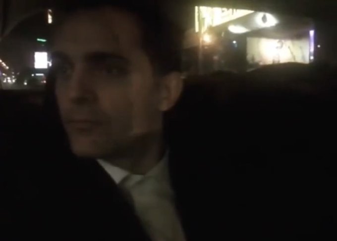 A man in a dark suit sits in the back of a car at night, illuminated by streetlights and signs visible through the window, as the post jokingly suggests he's "hearing things" after a long break from posting sol memes.