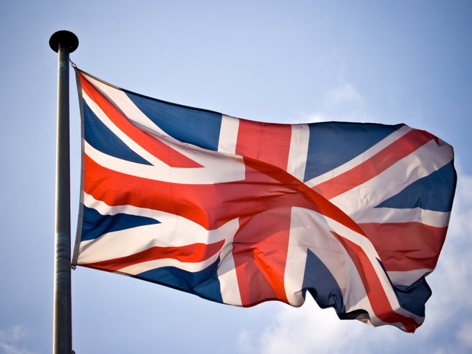 A Union Jack flag waves against a partly cloudy blue sky, mounted on a flagpole. The image symbolizes the United Kingdom, directly representing the country at the center of the article about its ambitions to launch tokenised sovereign bonds by 2027.