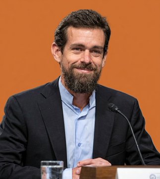 The image features a man wearing a suit, sitting behind a podium, and smiling for the camera. He has a beard and is seated in front of a microphone, suggesting that he might be giving a speech or presentation. The background is orange, which contrasts with his dark suit and adds visual interest to the scene. There are two cups placed on either side of him, possibly containing water or other beverages. The image also includes text that reads "Jack Dorsey: 'Choose to pay with Bitcoin at the counter'". This could be a reference to the man's recent speech about Bitcoin or another related topic.