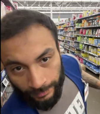 The image features a man with a beard wearing a blue shirt, standing inside a grocery store aisle filled with various products such as cereal boxes. The man appears to be making a funny face or expression while looking at the camera. There are multiple shelves of items in the background, including numerous cereal boxes and other food products.