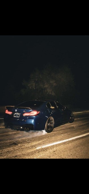 A dark blue BMW sedan drifting or burning rubber on a road at night, with smoke billowing from the rear tires and red taillights illuminated.